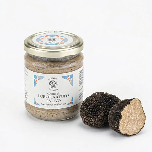 Uma fotografia close-up em luz natural de uma trufa negra de verão inteira e outra cortada ao meio, revelando o interior marmorizado pálido. Fatias finas e frescas de trufa repousam sobre uma mesa de madeira rústica ao lado de uma única flor roxa de açafrão com seus estigmas vermelhos, com um fundo de cozinha embaçado.