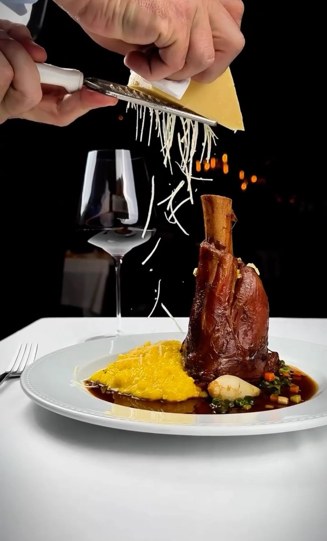 Pernil de cordeiro ou ossobuco suculento sobre uma base de polenta cremosa e vegetais, com molho escuro e um copo de vinho tinto ao fundo. Um chef está ralando queijo parmesão fresco por cima, realçando o prato em um ambiente sofisticado.