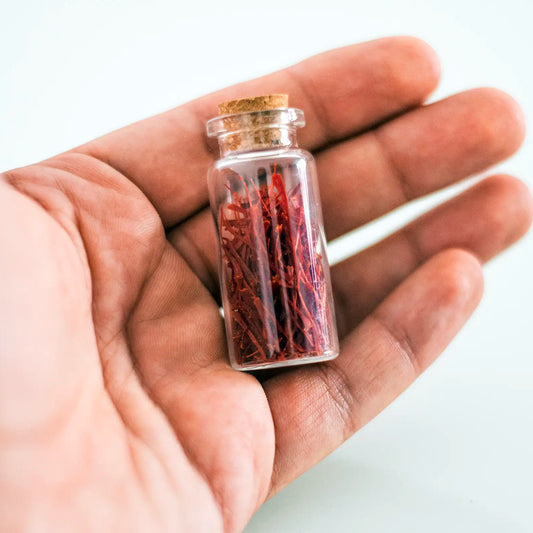 Mão segurando um pequeno frasco de vidro com tampa de cortiça, repleto de pistilos de açafrão de 1 grama da MA Saffron, em um close-up detalhado. A imagem foca na delicadeza do tempero e na facilidade de manuseio e degustação, transmitindo confiança na pureza e frescor do açafrão iraniano. Uma representação tangível da porção individual, ideal para cozinhas gourmet no Brasil, distribuída pela Pregiato.