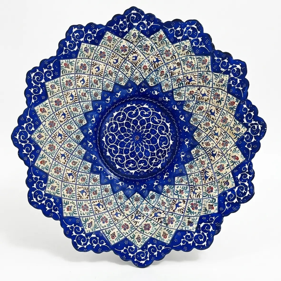 Uma vista de close-up de um único e magnífico prato redondo de metal, decorado com a técnica de esmaltação persa Minakari (میناکاری). O prato possui uma borda elegantemente recortada e uma pintura à mão intrincada com motivos geométricos e florais simétricos em tons intensos de azul-turquesa, azul-cobalto e branco, típicos da arte de Isfahan. Uma mão segura o prato contra um fundo desfocado de vegetação natural, destacando a peça como um item decorativo de luxo.
