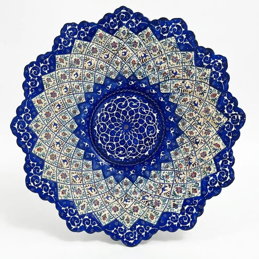 Uma vista de close-up de um único e magnífico prato redondo de metal, decorado com a técnica de esmaltação persa Minakari (میناکاری). O prato possui uma borda elegantemente recortada e uma pintura à mão intrincada com motivos geométricos e florais simétricos em tons intensos de azul-turquesa, azul-cobalto e branco, típicos da arte de Isfahan. Uma mão segura o prato contra um fundo desfocado de vegetação natural, destacando a peça como um item decorativo de luxo.