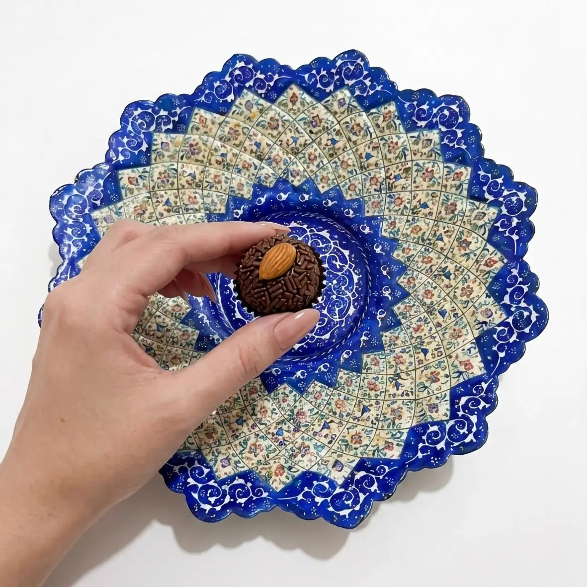 Vista frontal de um prato esmaltado iraniano com desenhos em azul cobalto, feito por um artista iraniano, mostrando uma mulher colocando um bragadeiro de chocolate brasileiro no prato.