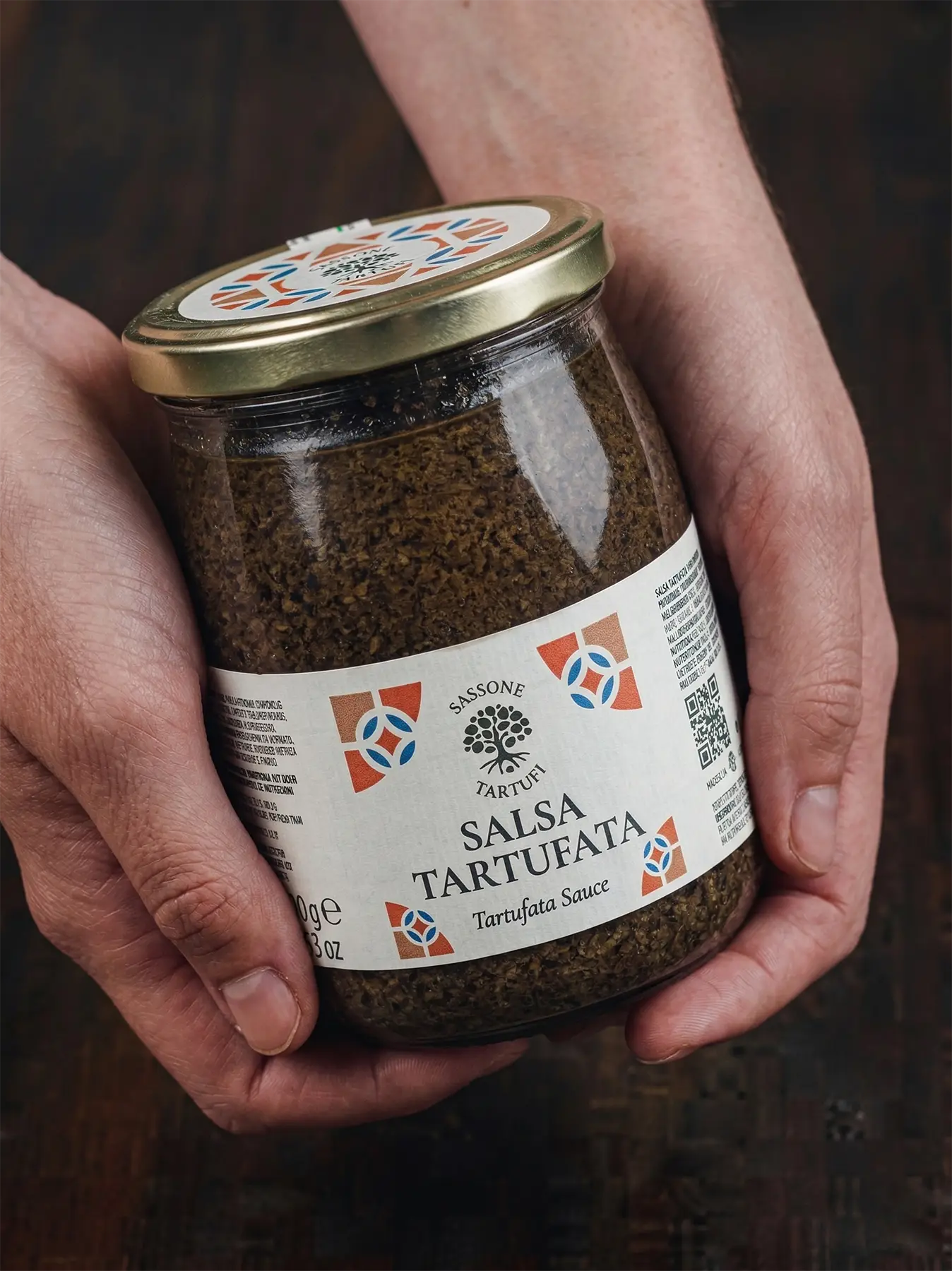 Mãos segurando um pote de salsa tartufata, destacando a textura rústica e a qualidade premium do autêntico molho de trufas da Itália.