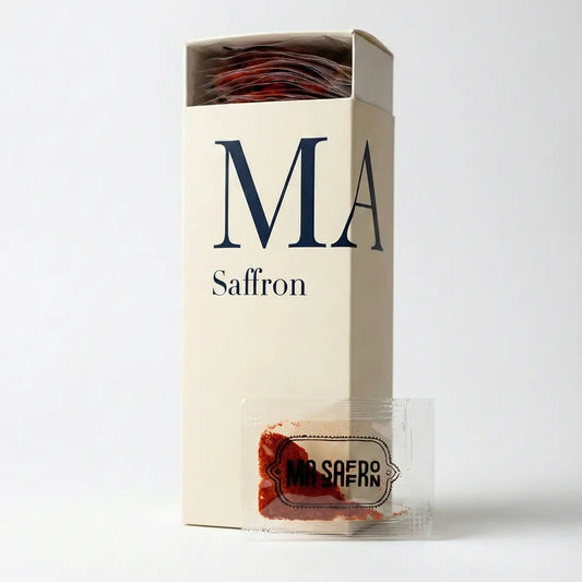 Caixa expositora da marca MA Saffron aberta, revelando múltiplos sachês individuais de açafrão iraniano puro. Em destaque, um sachê transparente mostrando a cor vibrante da especiaria. Formato prático e higiênico, ideal para uso culinário fracionado e revenda em empórios gourmet pela Pregiato.