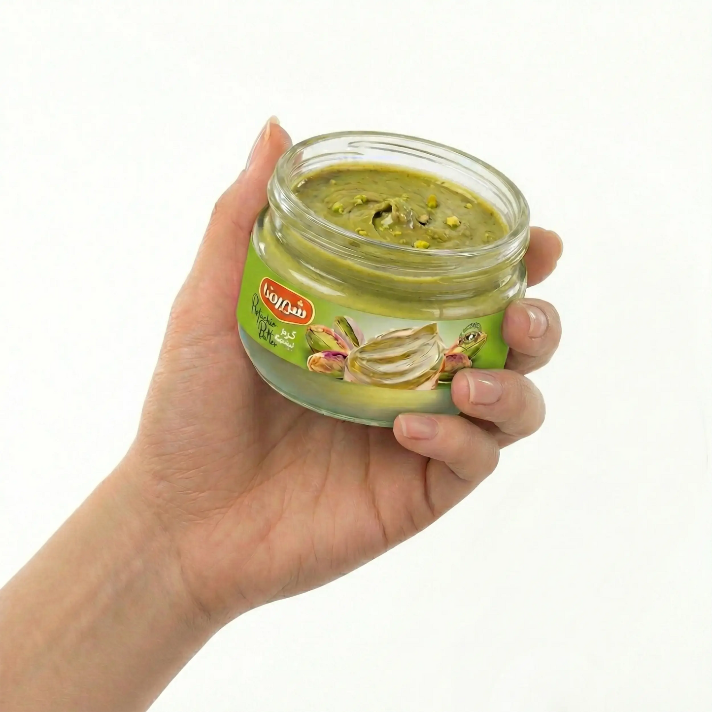Pote aberto de manteiga de pistache sendo segurado, revelando a textura espessa, verde e natural com pedaços reais de pistache (crunchy). Imagem que desperta o apetite e comprova a alta concentração de pistache puro no produto.